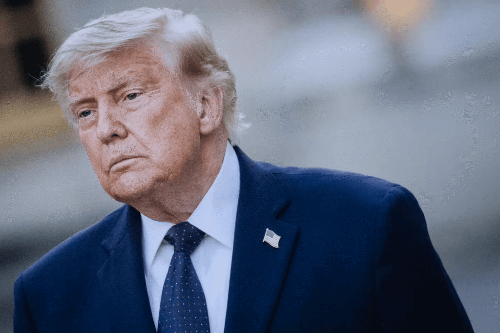 Donald Trump in a navy suit and blue polka-dot tie with an American flag lapel pin, looking serious outdoors.
